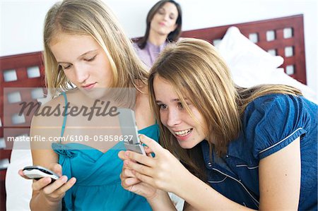 2 smiling teenage girls using mobile phones, woman in background