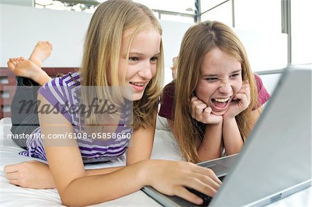 2 teen girls lying on bed using laptop