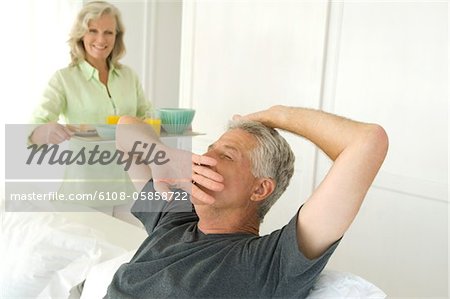 Woman bringing breakfast to man yawning in bed