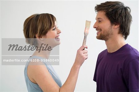 Young woman holding a paintbrush in front of a mid adult man's face