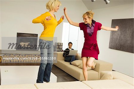 Two young women dancing with a young man using a laptop in the background