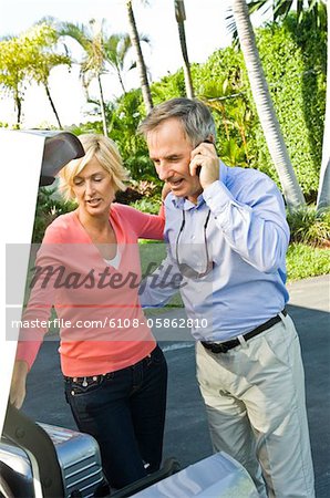 Man talking on a mobile phone beside his wife loading a luggage into car