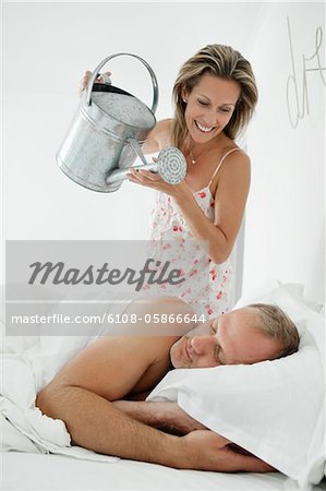 Woman pouring water with a watering can on a man sleeping on the bed