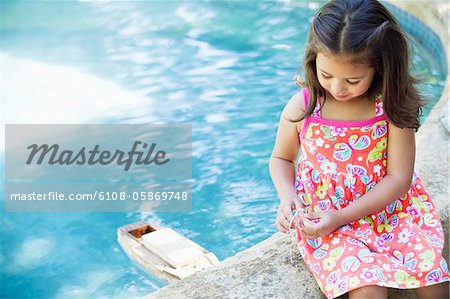 Girl sitting at edge of swimming pool looking at toy boat in water