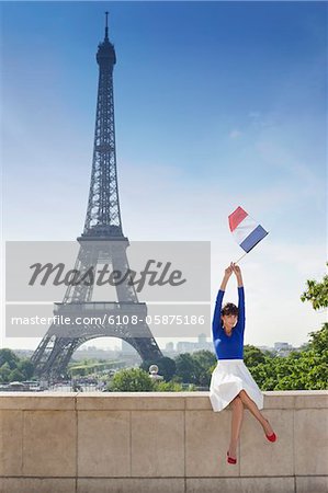 Woman holding a French flag sitting on a stone wall with the Eiffel Tower in the background, Paris, Ile-de-France, France