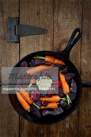 Raw root vegetables in a cast iron skillet ready for the oven and shot from above. Vegetables include carrots, red beets, garlic and purple potatoes.