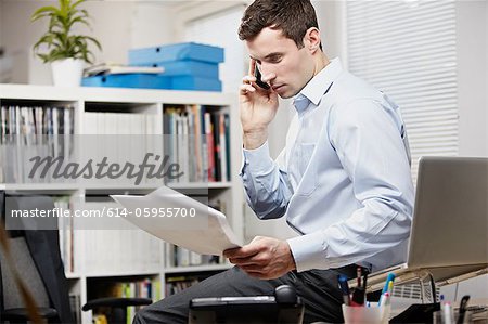 Büroangestellter sitzend am Schreibtisch, auf Handy und betrachten von Papierkram