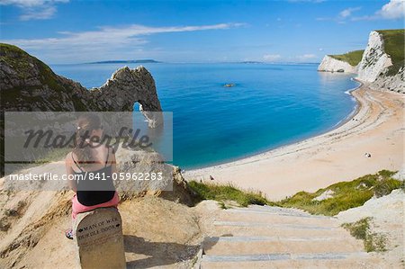 Woman resting on a clifftop sign, looking towards Durdle Door beach, Jurassic Coast, UNESCO World Heritage Site, Dorset, England, United Kingdom, Europe