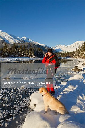 Skieur nordique s'arrête de répondre à son téléphone cellulaire alors que son chien est assis à côté de lui, la rivière Mendenhall, sud-est de l'Alaska, Juneau, hiver