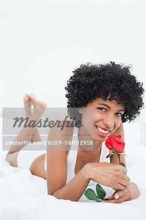 Portrait of a smiling black haired woman smelling a rose while lying on her bed