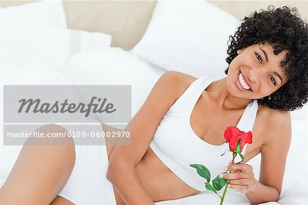 Portrait d'une femme souriante tenant une rose sur son lit dans une chambre lumineuse
