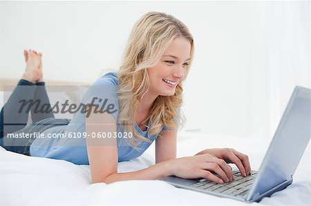 Une femme est couchée sur le lit souriant et son ordinateur portable est en face d'elle.