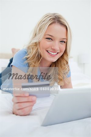 A woman lying on the bed with her tablet and credit card, smiling and looking straight ahead.