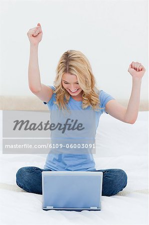 A woman is celebrating with arms in the air as she looks at her laptop while sitting on the bed.