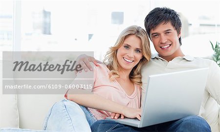 A man and woman sit on the couch together smiling as they use the laptop and look in front of them.