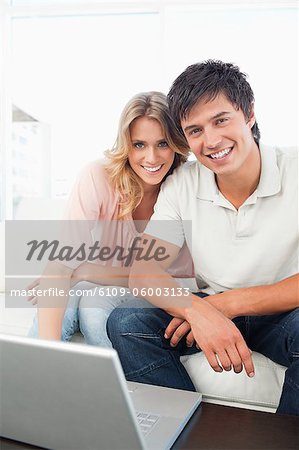 A man and woman sitting on the couch with a laptop in front of them, as they look in straight ahead and smile