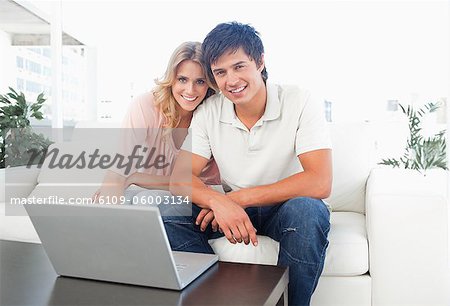 A man and woman sitting on the couch together, smiling  with the laptop on a table in front of them