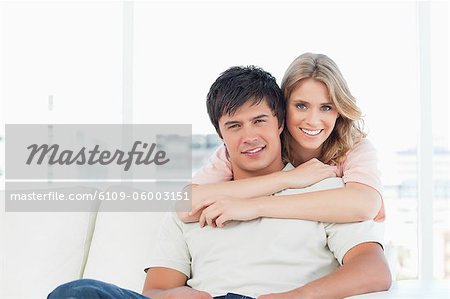 Ein Mann sitzt auf der Couch mit der Frau hinter ihm stehend mit ihre Arme um ihn, wie sie Lächeln.
