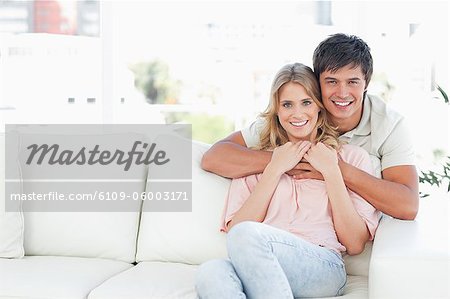 A man holds the woman from behind the couch as they both smile and look in front of them