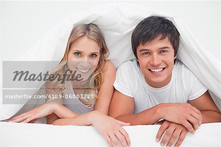 A man and woman beside each other at the edge of the bed smiling.