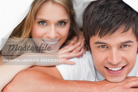 A close up shot, of a man and woman resting on their hands, under the quilt and smiling.