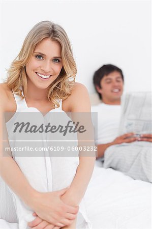 A close up shot of the woman sitting at the bottom of the bed, with the man at the top reading and both are smiling.