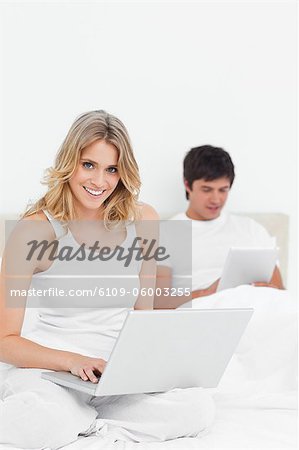 Ein Mann mit dem Tablet und Frau mit den Laptop sind beide Lächeln, beim Sitzen auf dem Bett.