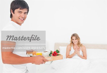 The man brings the woman breakfast in bed while both are smiling.