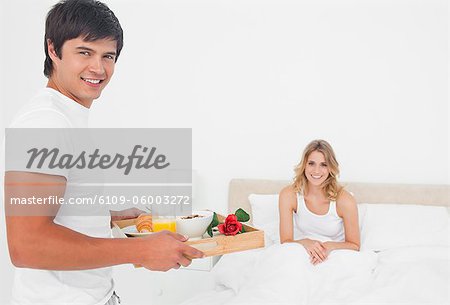 The man brings the woman some breakfast in bed, and both are smiling.