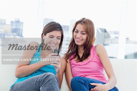 Teenager sitting on a sofa and sending a text while her friend is looking at her phone