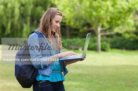 Young girl typing on her laptop while standing in a park and carrying a backpack