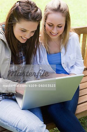 Young smiling teenagers looking at a laptop while sitting on a bench in a parkland