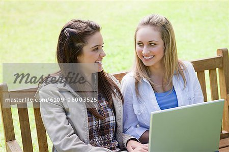 Smiling women sitting on a bench in the countryside with a laptop on their knees