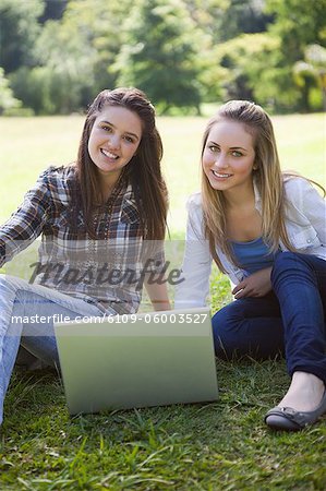 Young smiling girls sitting on the grass with a laptop while looking at the camera