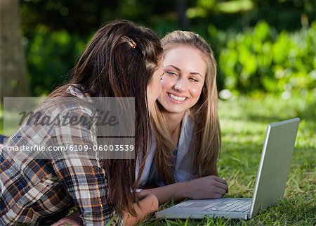 Young smiling students lying on the grass in a park while working with a laptop