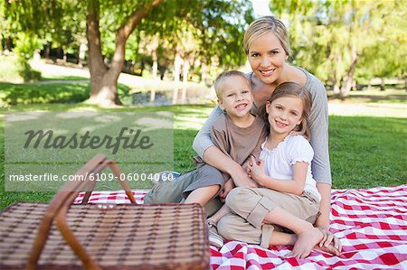 A mother her son and daughter all smile while sitting on a blanket in the park