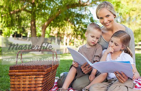 A smiling mother looks at the camera while her kids read the book she is holding