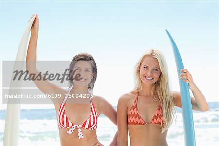 Two women smiling as they stand in bikinis while holding their perched surfboards on the beach