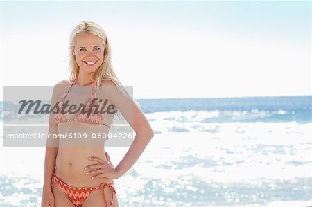 Woman in bikini smiling as she has one hand on her hip while she stands by the sea