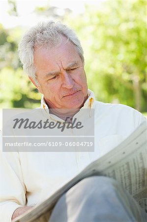 Man with a serious look on his face while looking at a newspaper as he sits on a bench in the park