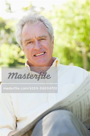 Man smiles as he holds a newspaper while sitting on a bench in the park