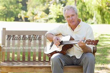 Homme regardant devant lui, alors qu'il joue une chanson sur une guitare, assis sur un banc dans le parc