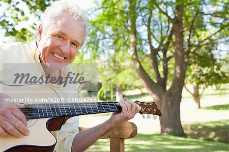 Man smiling while playing a guitar while he sirs on a bench with a tree in the background