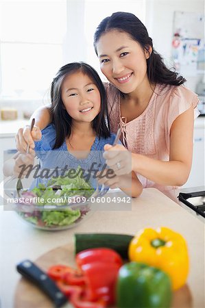 A smiling mother and daughter look ahead as they toss a salad together