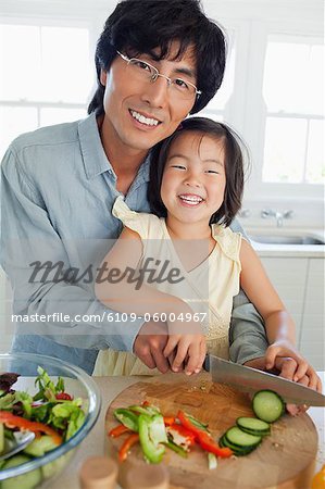 A smiling father and daughter cutting cucumbers together in the kitchen and looking forward
