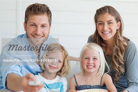 The smiling family sit on the couch together as the dad changes the tv channel