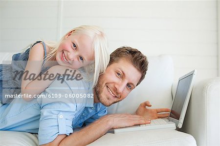 A father lies across the couch using his laptop as his daughter lies on top of him