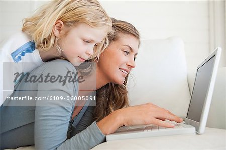 A smiling woman lies on the couch and interacts with her laptop as her daughter lies on top of her