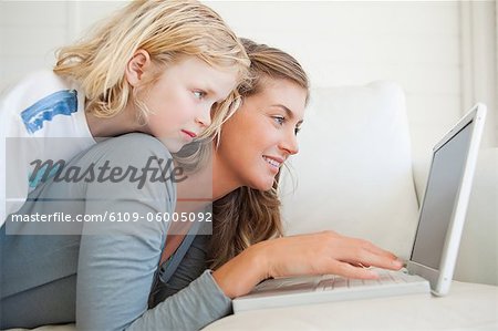 A child lies up on top of her mother as she uses the laptop on the couch and smiles