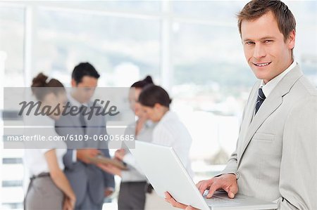 Smiling young salesman with his laptop and colleagues behind him